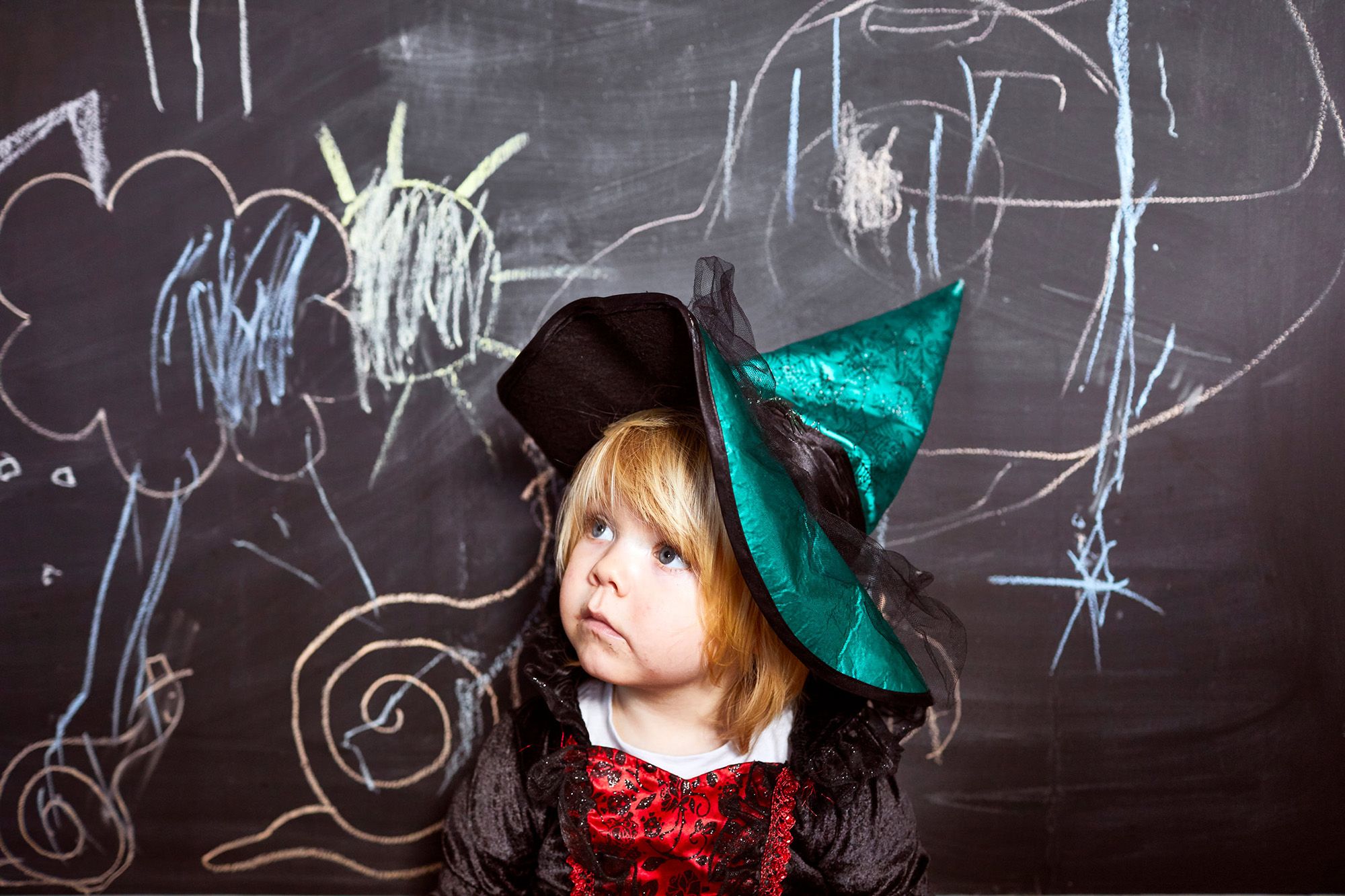 A young girl dressed as a witch, wearing a black and red dress and a pointed hat, standing in front of a blackboard with colorful drawings.