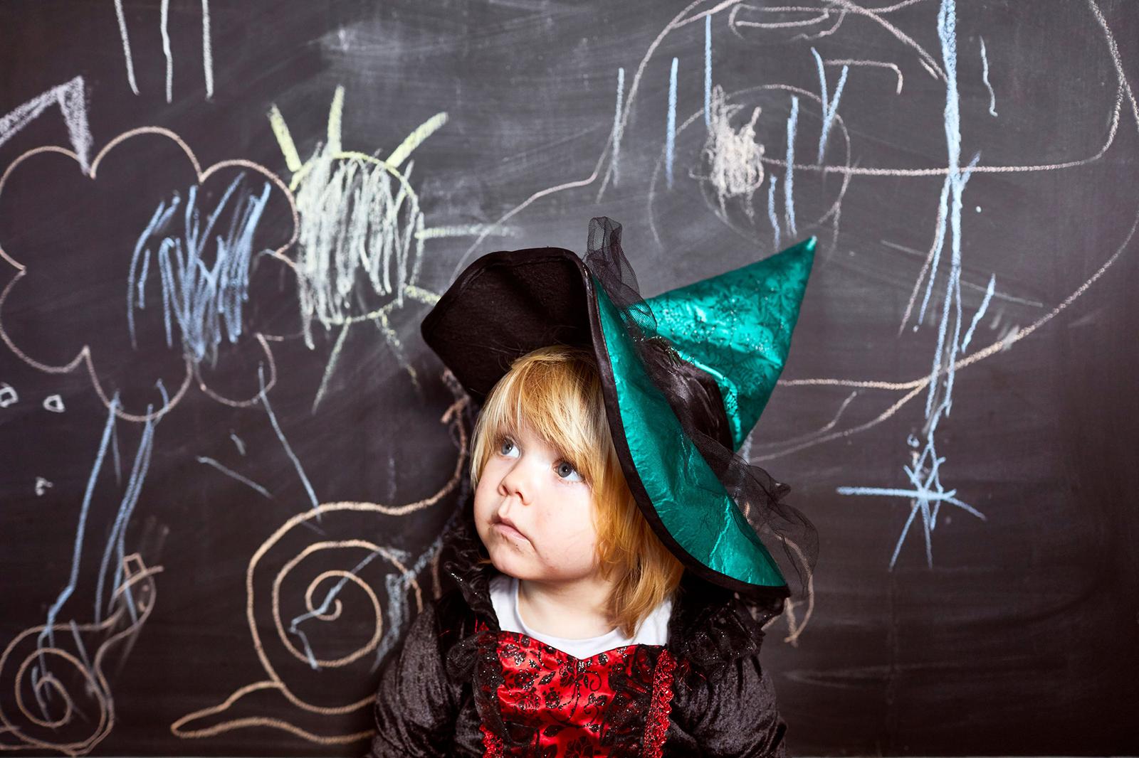 A young girl dressed as a witch, wearing a black and red dress and a pointed hat, standing in front of a blackboard with colorful drawings.