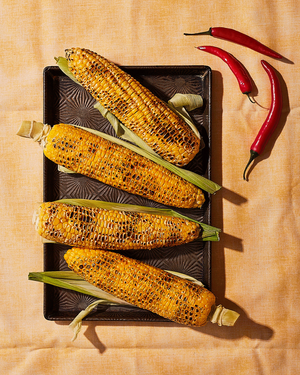 A still life of four grilled corn on the cob on a dark, ornate plate, with two red chili peppers in the top right corner.