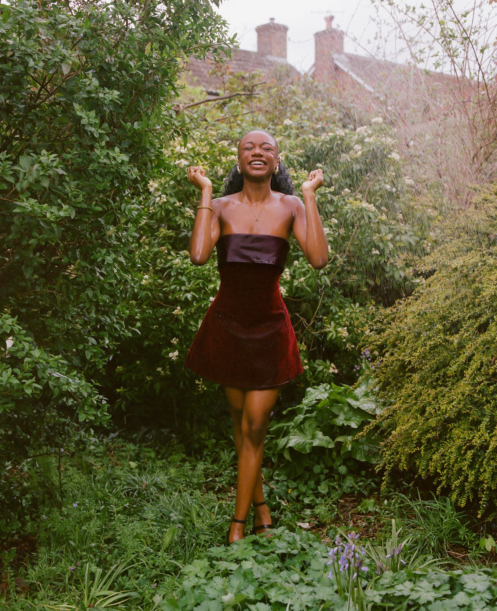 A woman in a red velvet mini-dress and black heels is standing in a lush, green garden, surrounded by trees and flowers, with a large, old-fashioned house in the background.