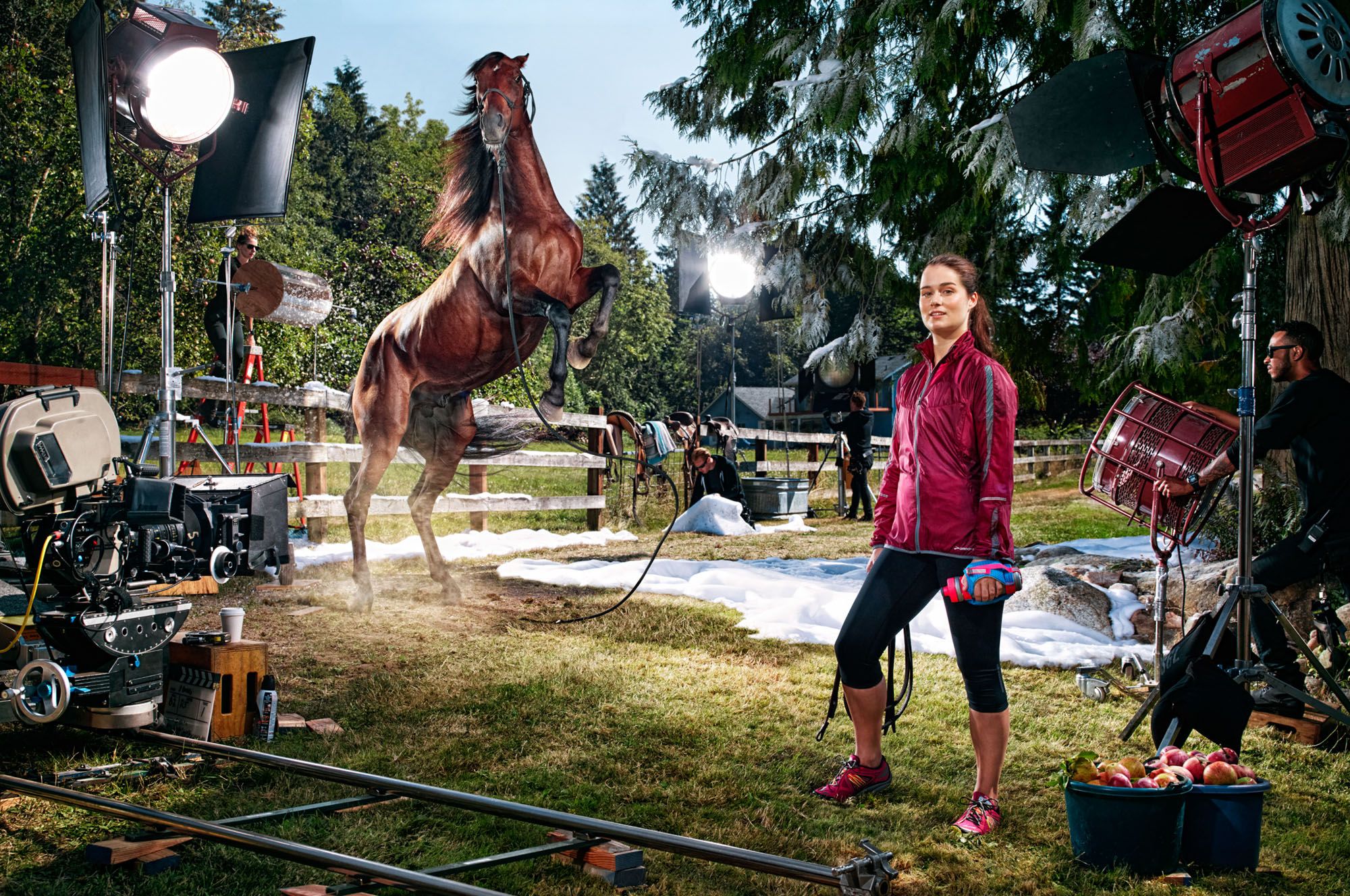 A woman in a red jacket and black leggings standing on a film set, with a horse in the background, surrounded by film equipment and a large tree in the distance.