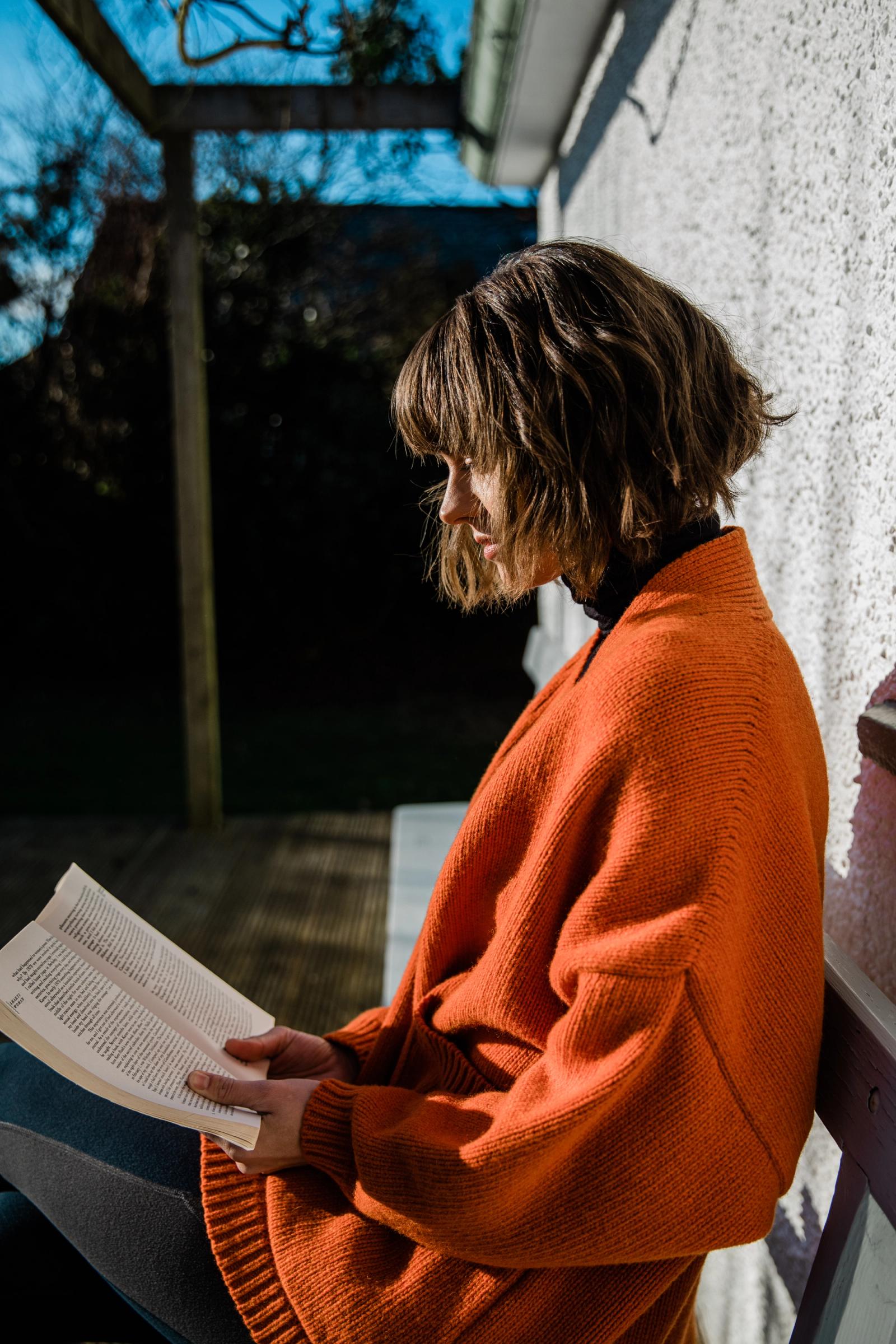 A woman in an orange sweater is sitting on a chair, reading a book.