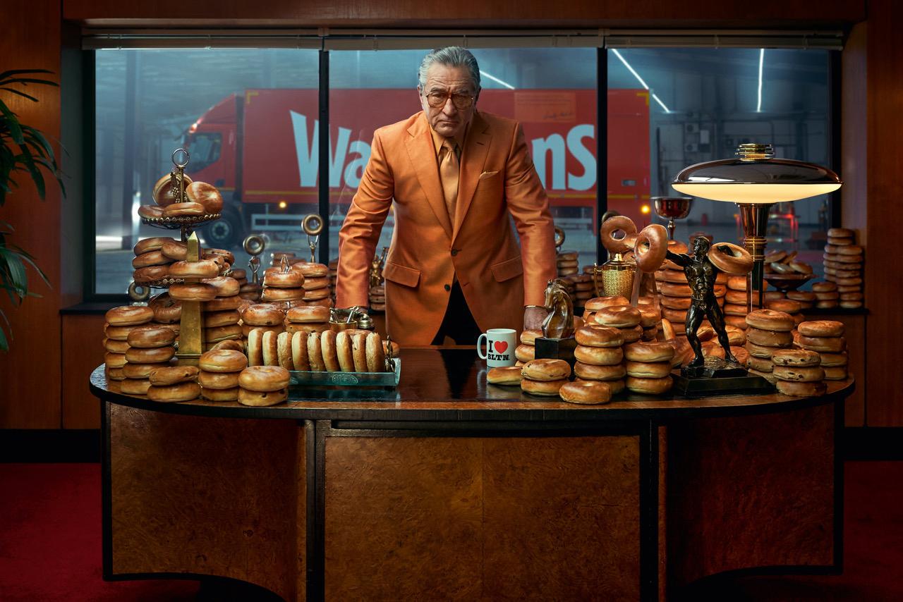A man in a bright orange suit standing behind a large desk covered in stacks of bagels, with a city street visible through the window behind him.