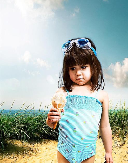 A young girl in a blue and white striped swimsuit, holding an ice cream cone, standing on a beach with tall grass in the background.