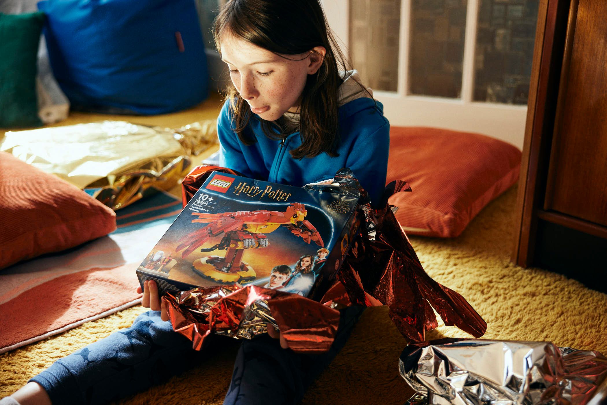 A young girl sits on a carpeted floor, holding a Harry Potter Lego set in her hands. She looks down at the box with a mixture of excitement and concentration, her face.