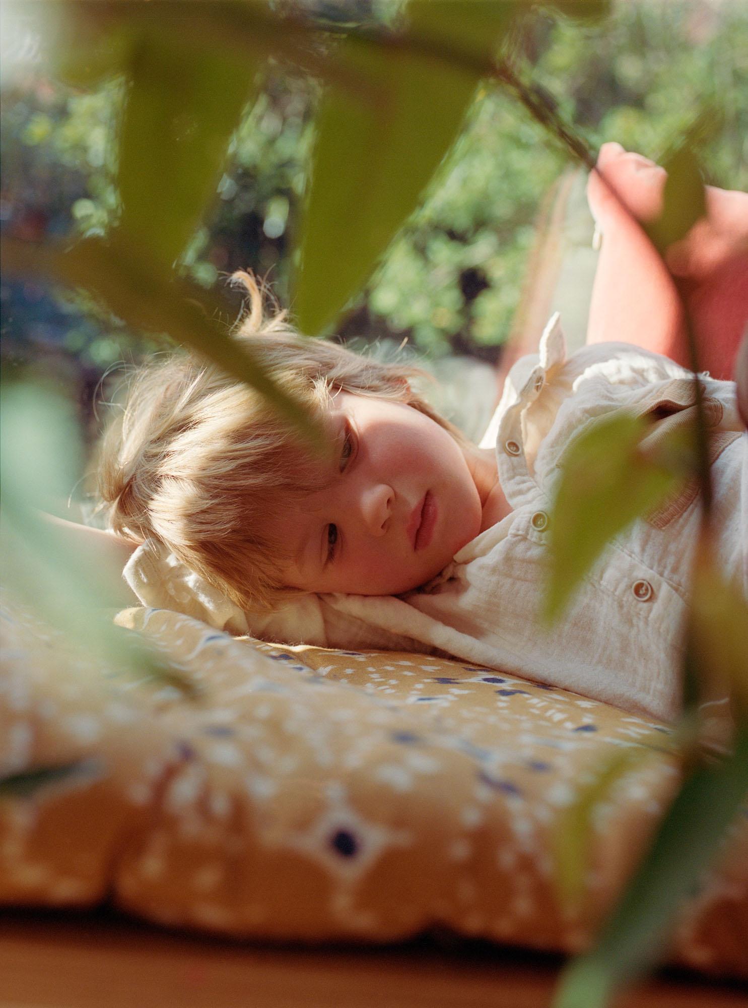 A young girl with blonde hair and a white shirt lying on a bed, surrounded by greenery.