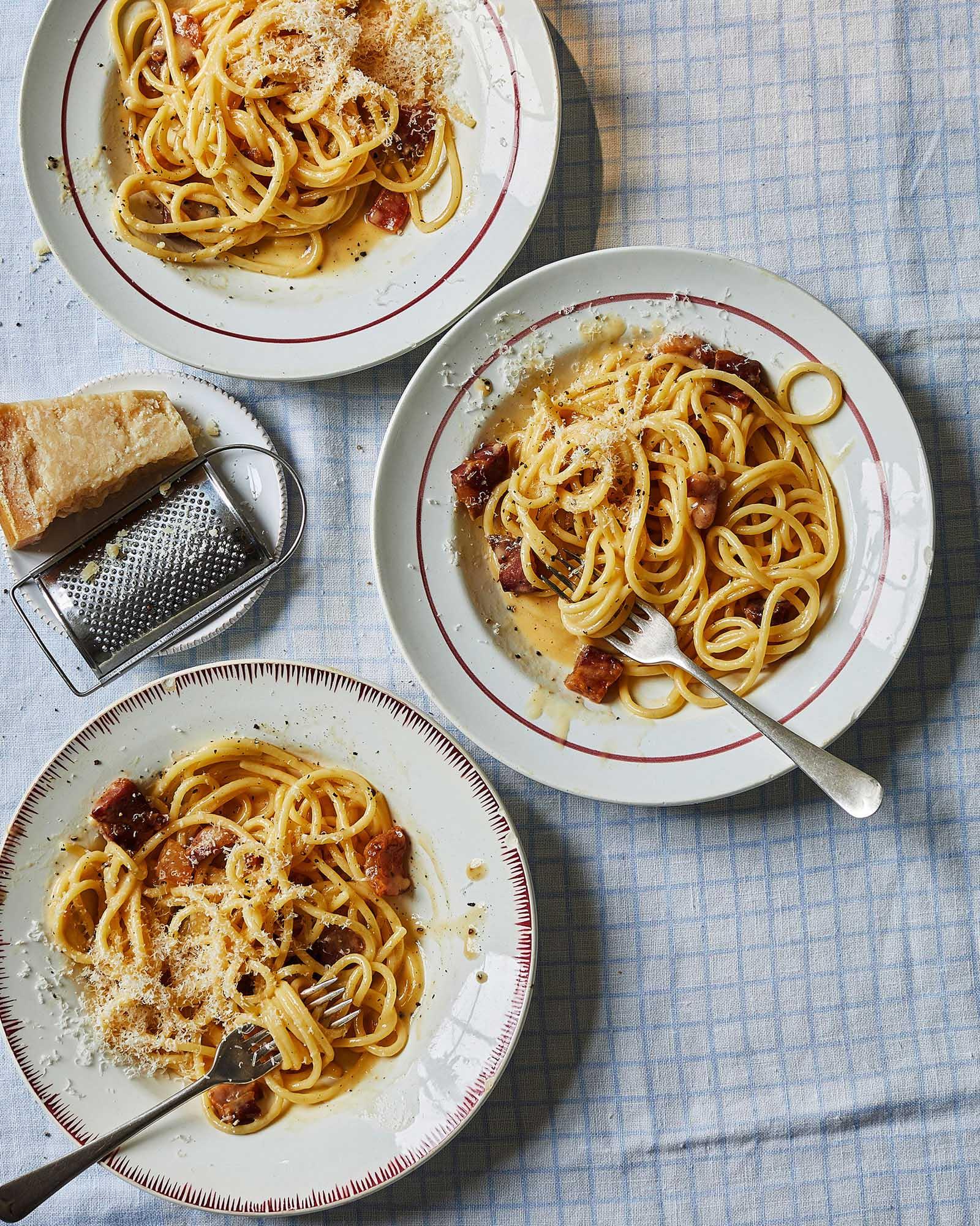 A table setting with three plates of pasta, a grater, and a loaf of bread.