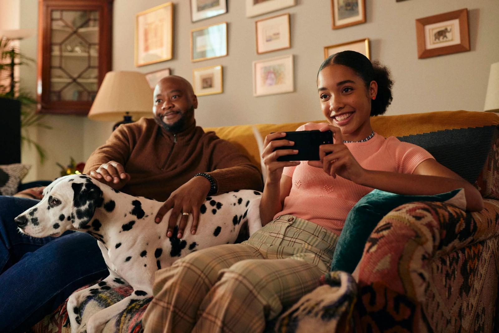 A man and a woman are sitting on a couch, with the woman holding a phone and the man petting a Dalmatian dog. The woman is wearing a pink top and plaid pants, while the man is.