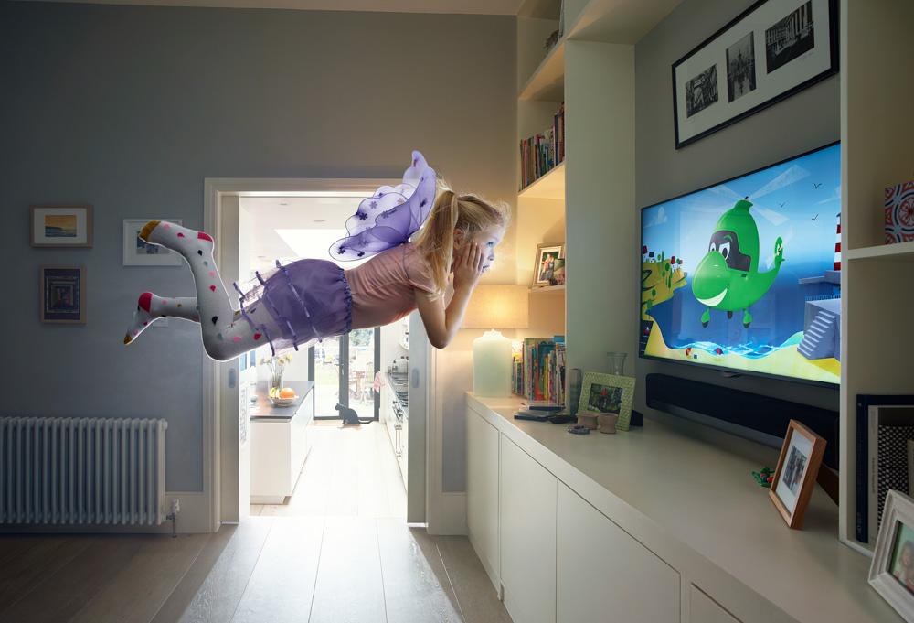 A young girl in a pink dress and purple fairy wings is flying through a living room, watching a cartoon on TV.