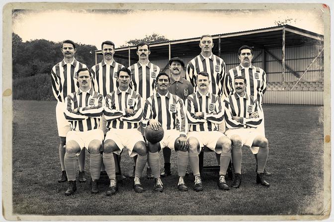 A group of men in football jerseys and shorts, posing in a field with a football in the foreground.