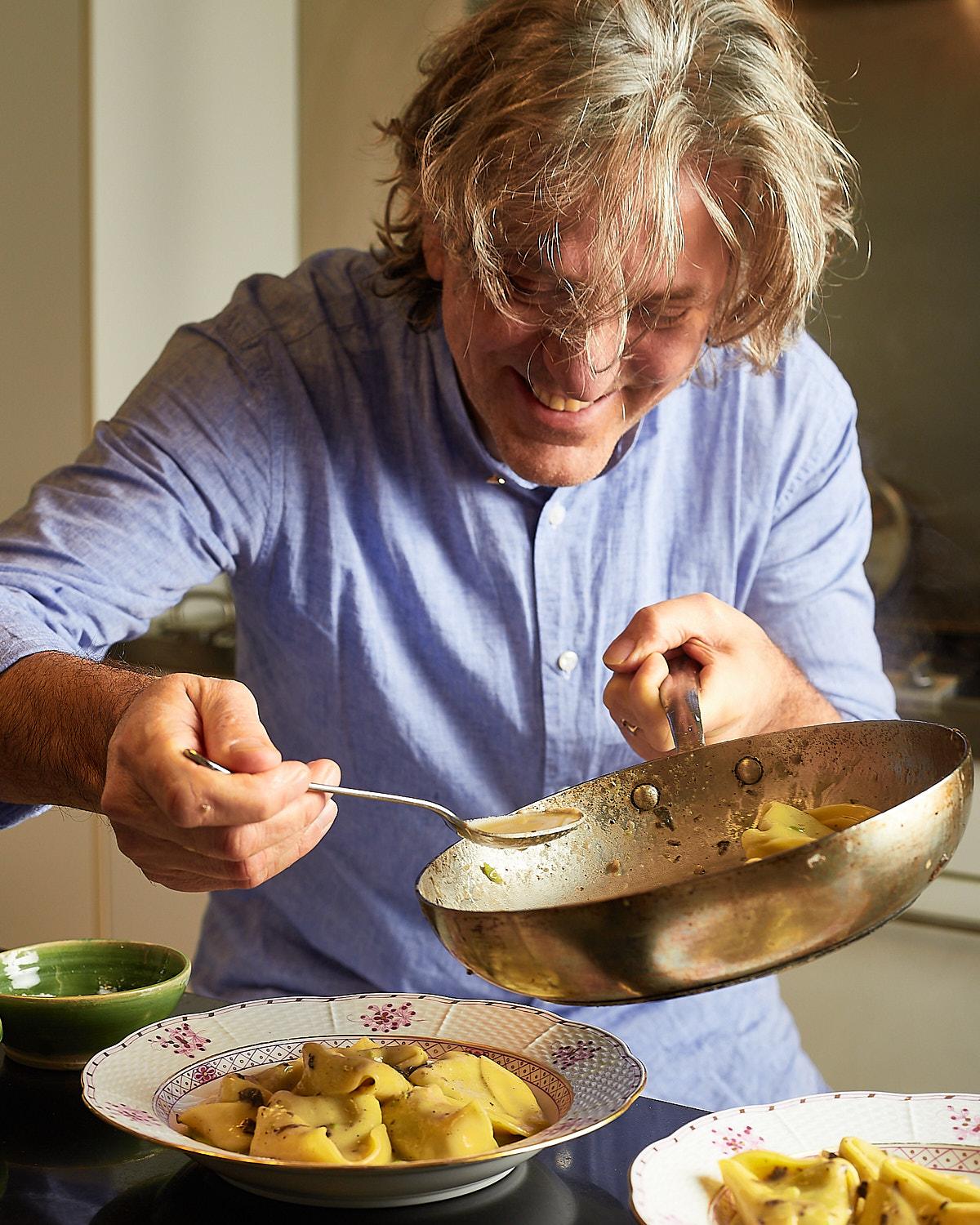 A man with messy, grey hair and a blue shirt is stirring a large metal pan with a spoon, with a bowl of pasta in the foreground.