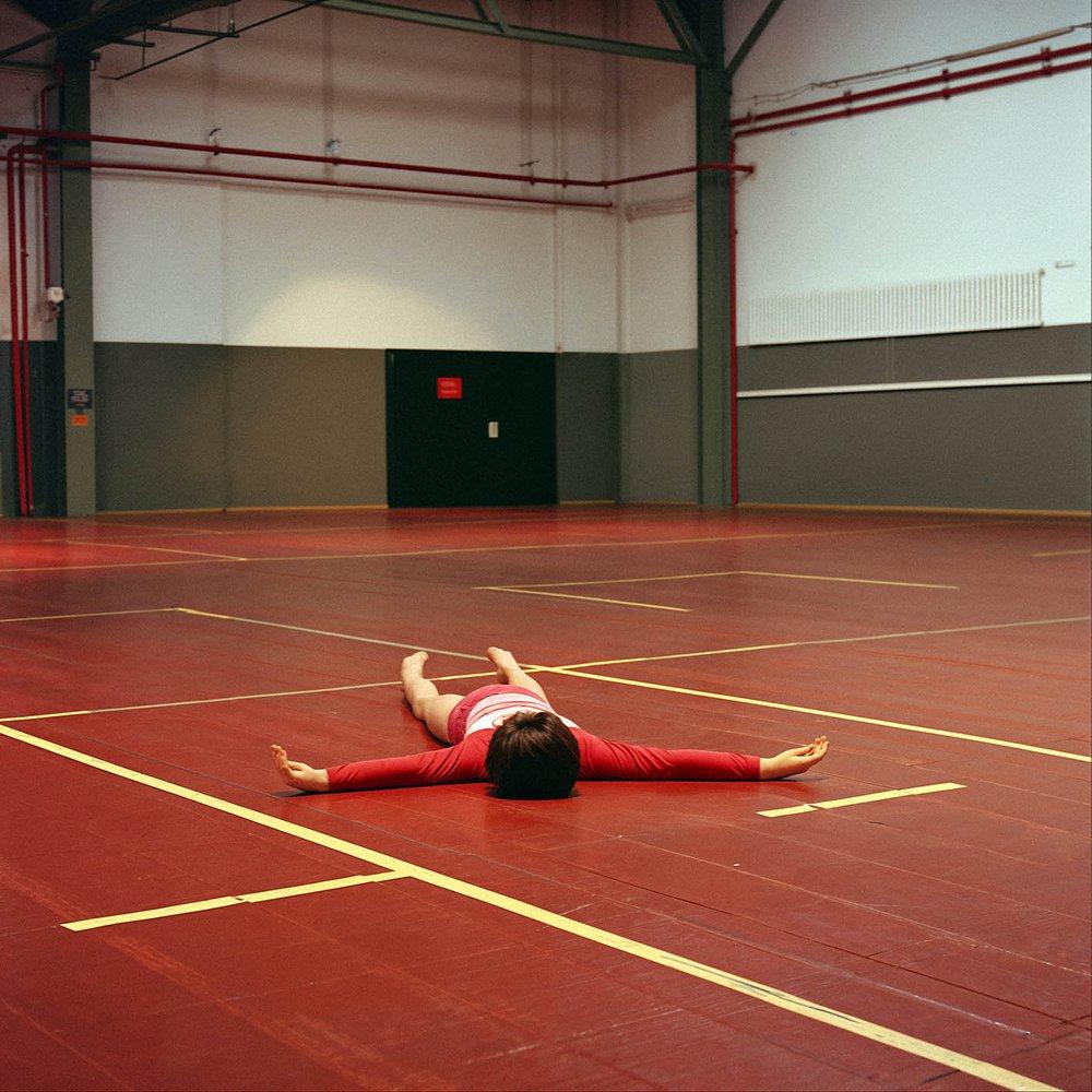 A person in a red and white striped leotard is lying on the floor in a large, empty, industrial-style room with a high ceiling and concrete walls.
