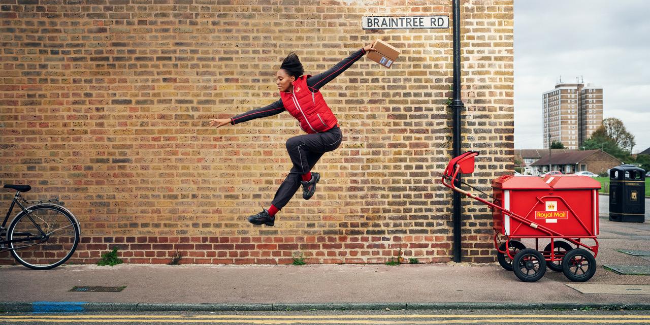 A man in a red and black jacket and black pants is jumping in the air, with a red mail cart and a brick wall in the background.