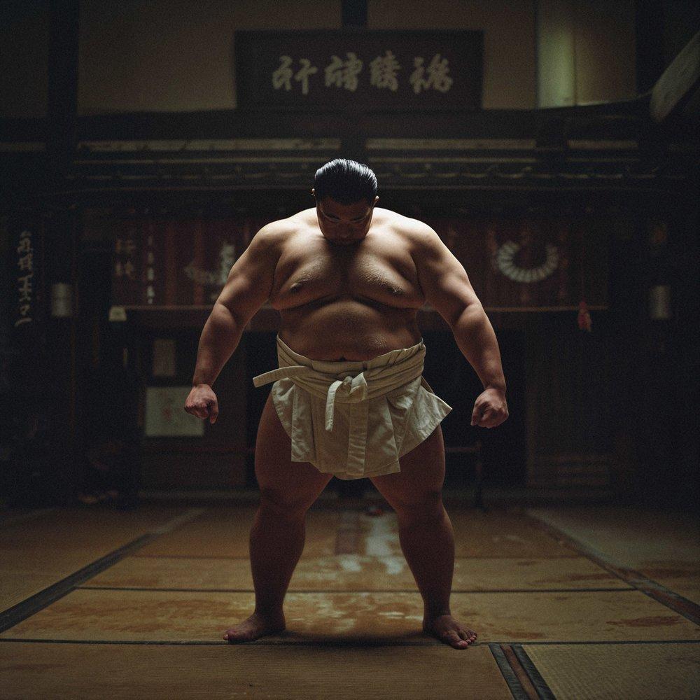 A sumo wrestler in traditional mawashi standing in a dimly lit dojyo.