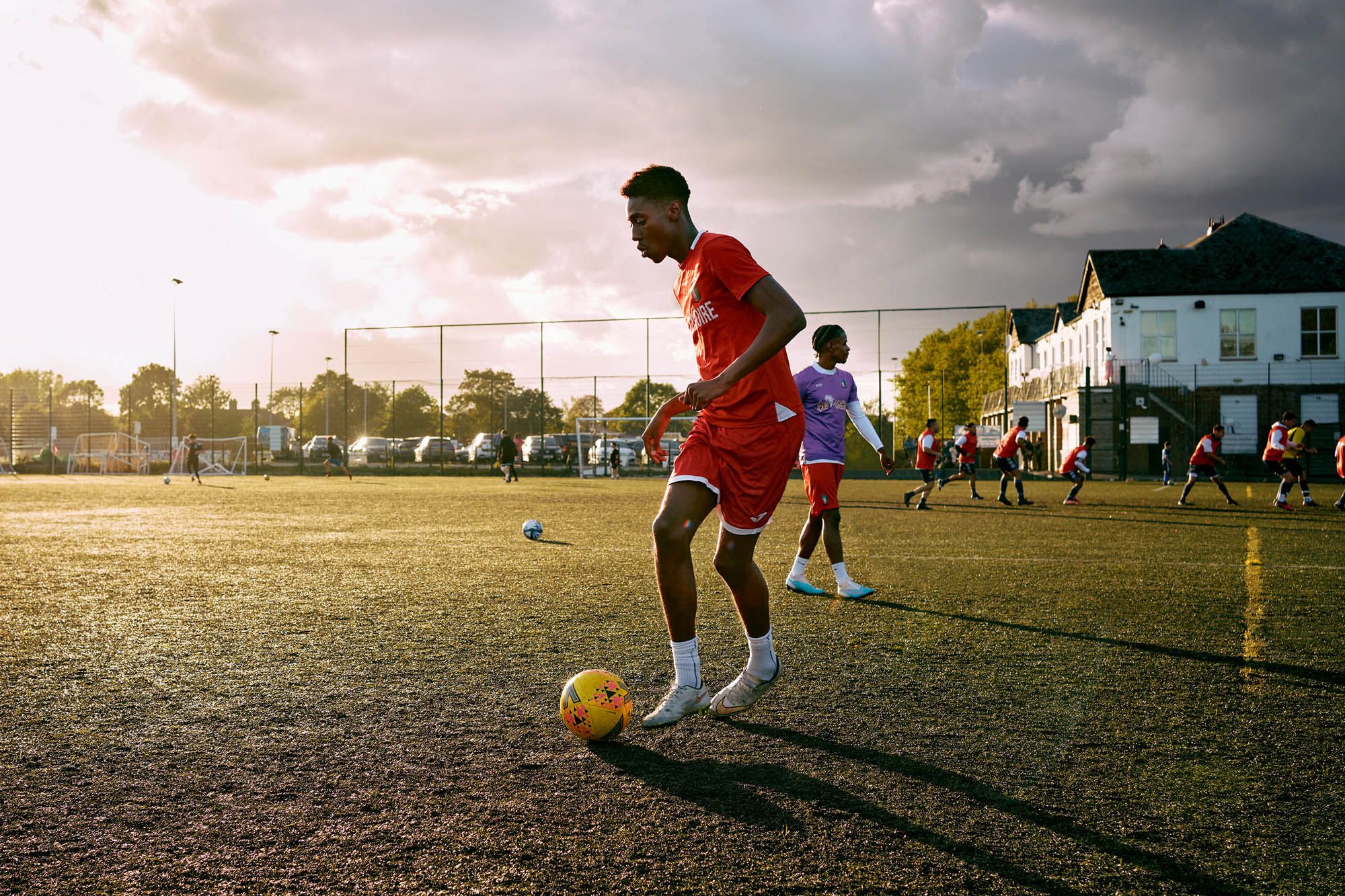 A group of young people, likely teenagers, are playing soccer on a field, wearing red and purple jerseys and shorts, with a few balls scattered around them.