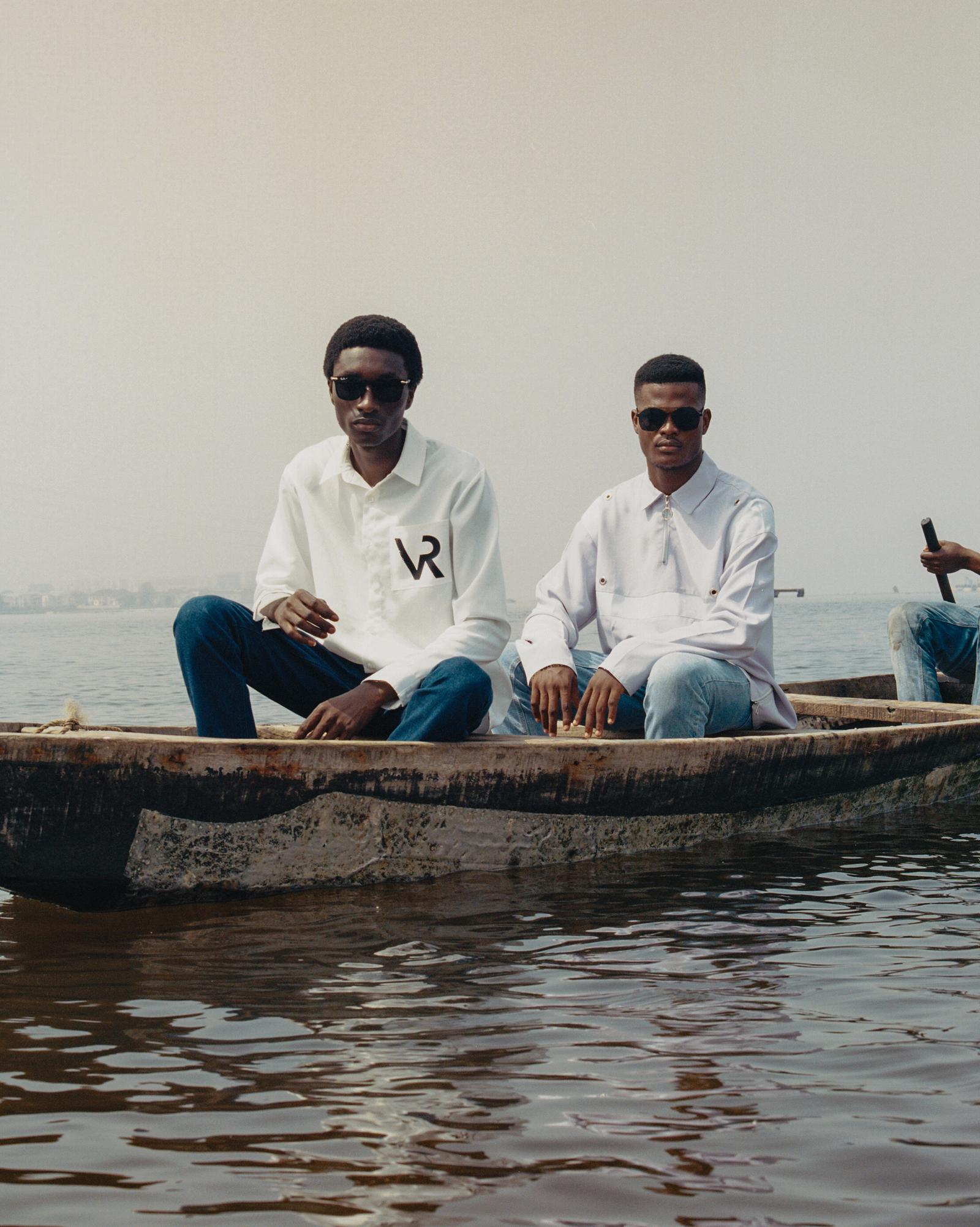 Two men in white shirts and jeans sitting in a wooden boat on a body of water.