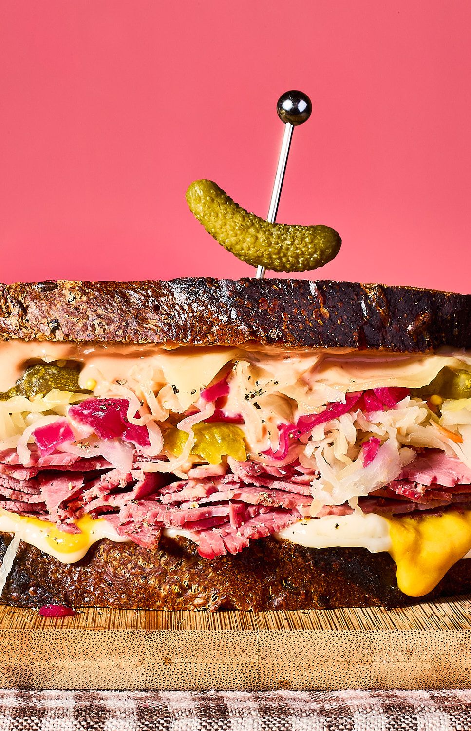 The image shows a towering Reuben-style sandwich on dark rye bread against a solid pink background. It is packed with layers of thinly sliced corned beef, sauerkraut, melted cheese, and Russian dressing, topped with a single cocktail gherkin held by a silver pick.