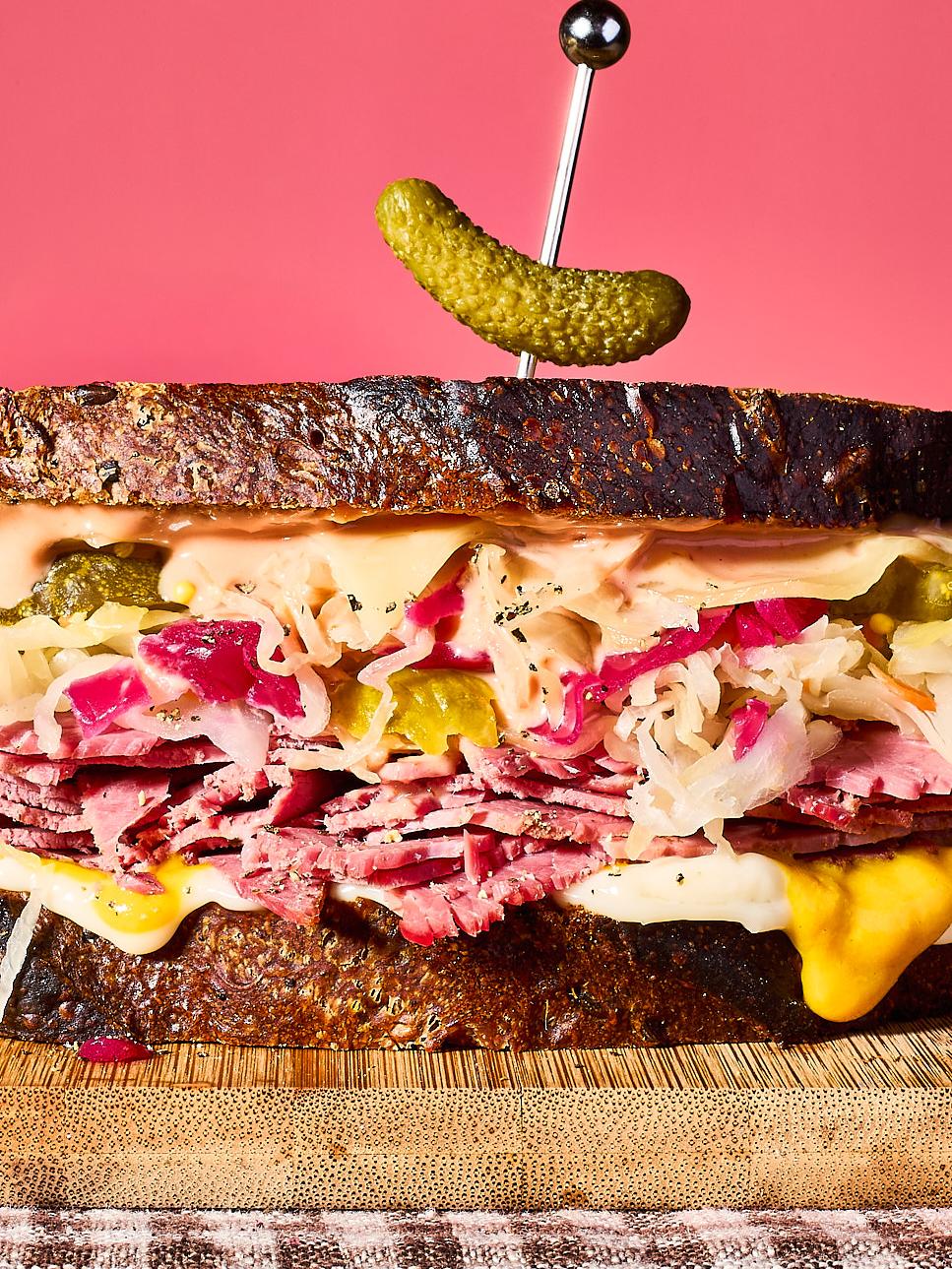 The image shows a towering Reuben-style sandwich on dark rye bread against a solid pink background. It is packed with layers of thinly sliced corned beef, sauerkraut, melted cheese, and Russian dressing, topped with a single cocktail gherkin held by a silver pick.