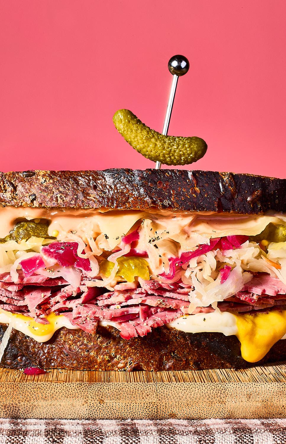 The image shows a towering Reuben-style sandwich on dark rye bread against a solid pink background. It is packed with layers of thinly sliced corned beef, sauerkraut, melted cheese, and Russian dressing, topped with a single cocktail gherkin held by a silver pick.