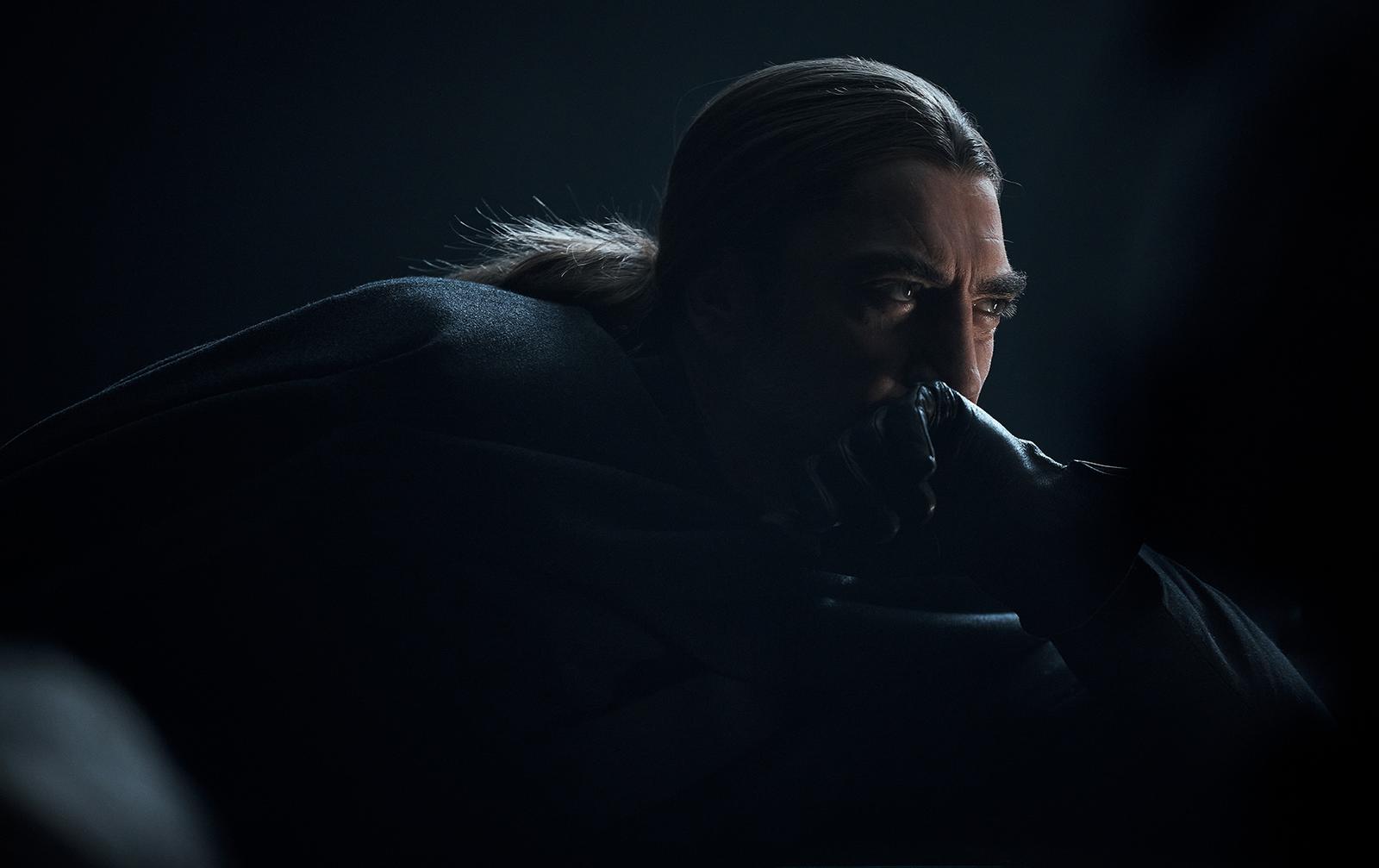 Javier Bardem, a man with long, dark hair and a beard, wearing a black leather jacket and a white shirt, looking directly at the camera with a serious expression.