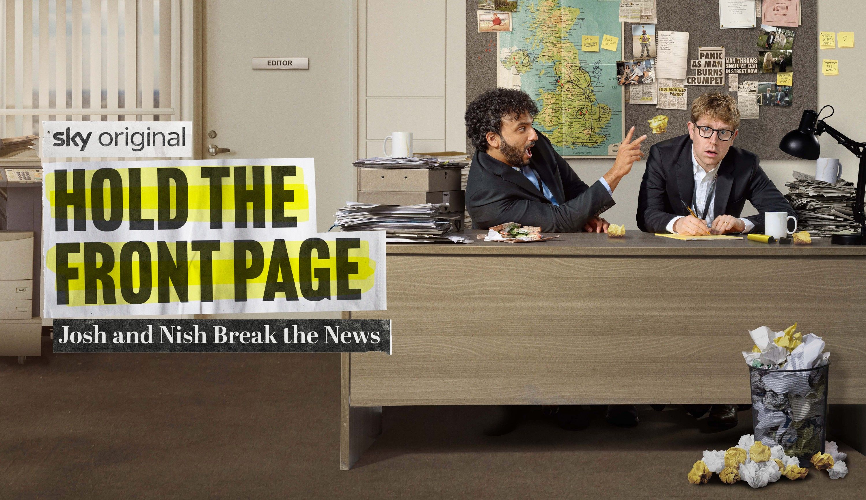 Two men in a newsroom, one holding a piece of food, sitting at a desk with a map of the UK on the wall behind them.