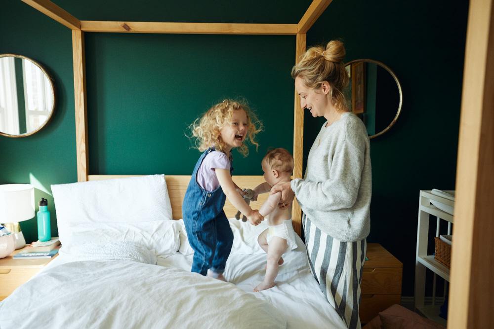 A young blonde mother, wearing a grey sweater and striped pants, stands in a bedroom with a dark green wall, holding a baby in one arm and a toddler in the other. The toddler is.
