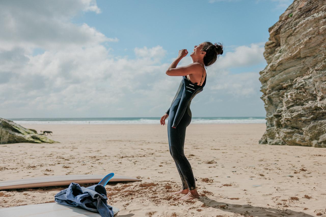 A woman in a wetsuit standing on a beach, with a surfboard and a towel in the background.