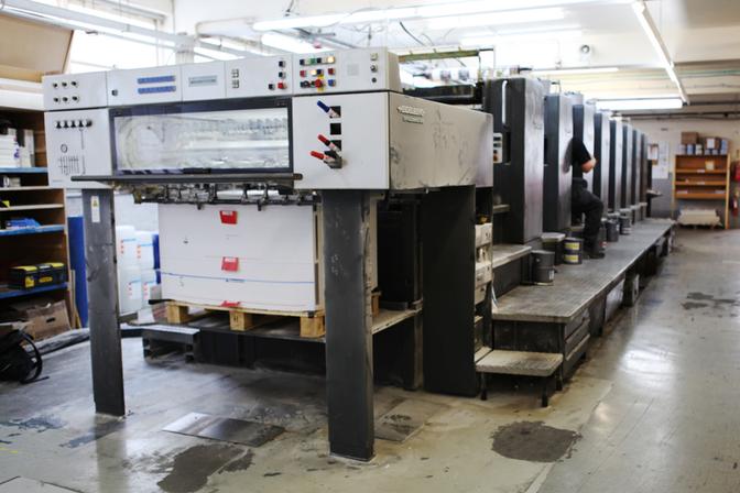 A large, industrial printing press in a warehouse or factory setting, with a worker in the background.
