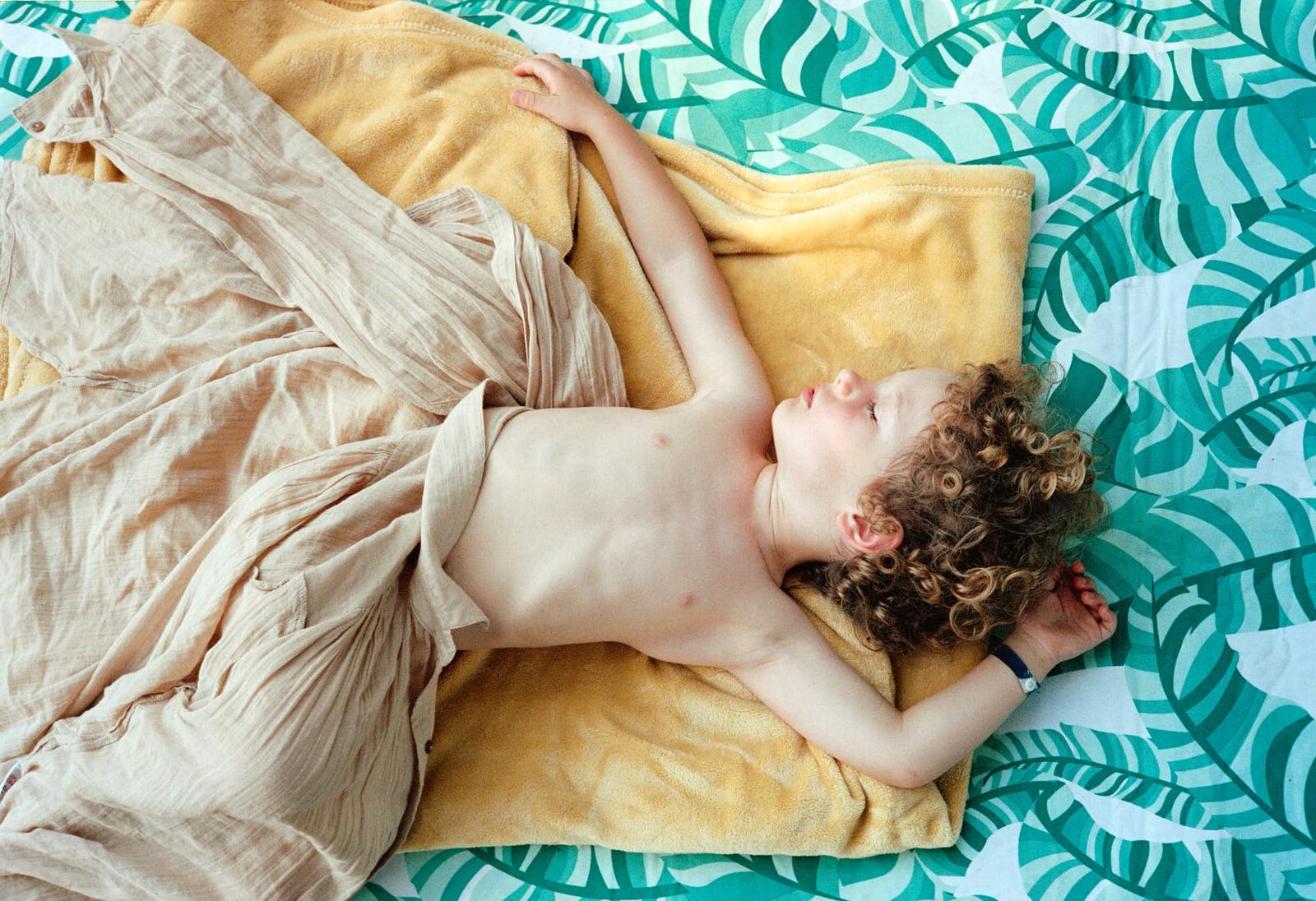 A sleeping child, barefoot and covered in a light brown blanket, lies on a bed with a vibrant green and white leaf-patterned sheet. The child's hair is a messy, curly brown, and.