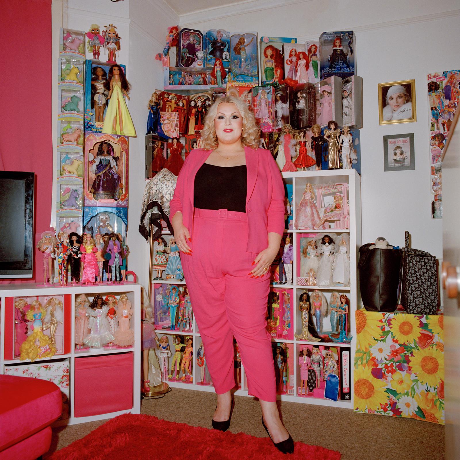 A woman stands in front of a wall displaying Barbie dolls and other collectible toys.
