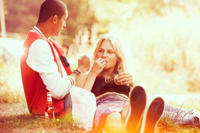 A young woman with long blonde hair is sitting on a blanket in a park, eating a sandwich and drinking a beer with a young man wearing a red and white striped rugby jersey.