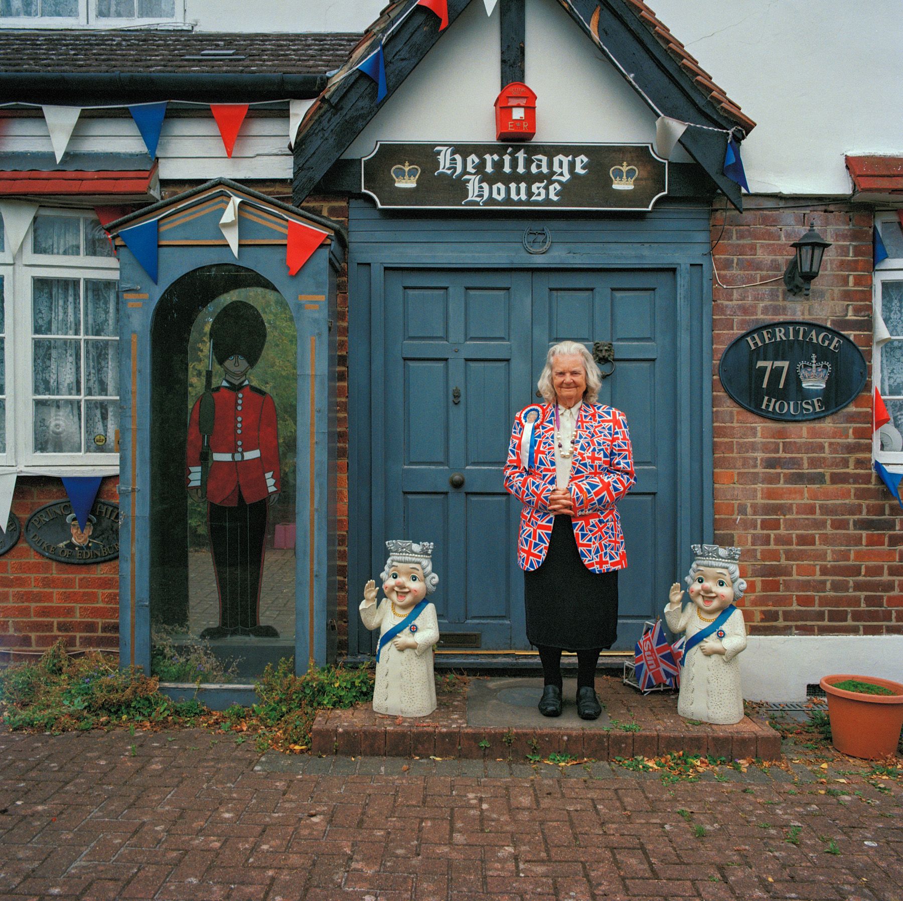 A woman stands in front of a heritage house holding a Union Jack flag.