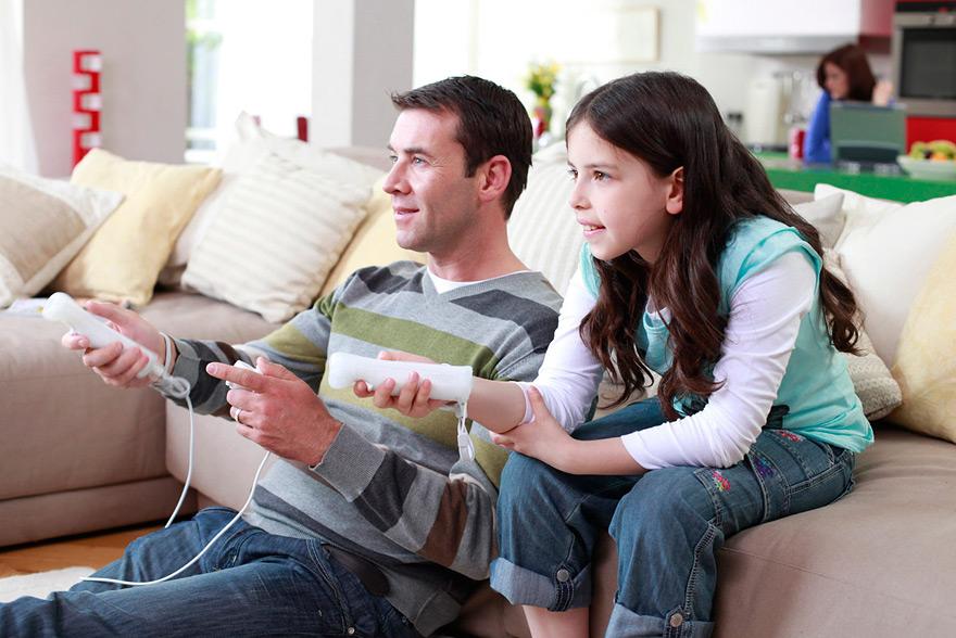 A man and a young girl sitting on a couch, both holding Nintendo Wii controllers and playing a video game together.