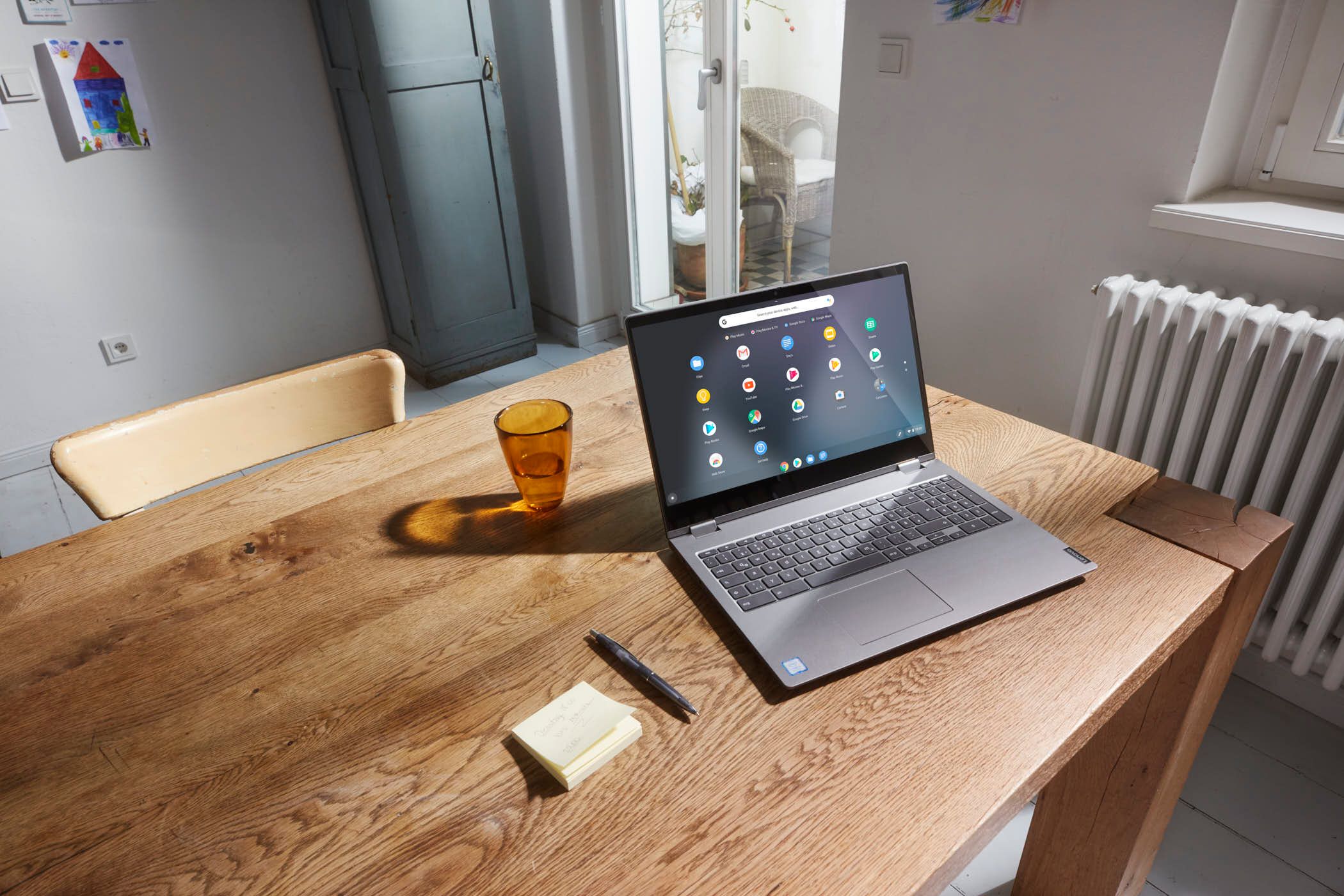 A silver laptop sits on a wooden table in a cozy, sunlit room. The screen displays the Google Chrome browser, and a stack of sticky notes and a pen are nearby. The room has white.
