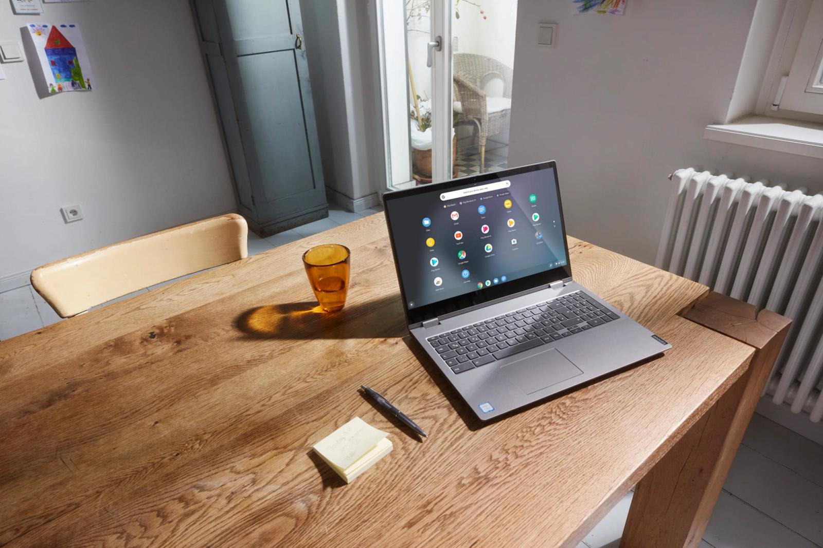 A silver laptop sits on a wooden table in a cozy, sunlit room. The screen displays the Google Chrome browser, and a stack of sticky notes and a pen are nearby. The room has white.