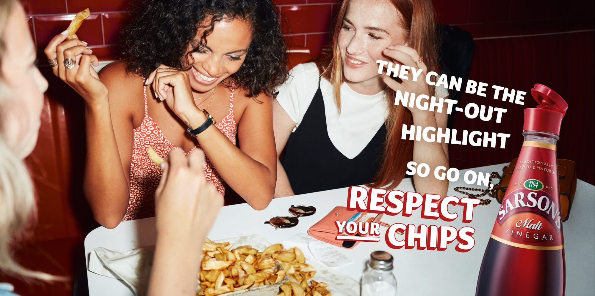 Two women are eating fried food at a table. The woman in the foreground is smiling and holding up a fried food item. The woman in the background is looking at her and smiling..