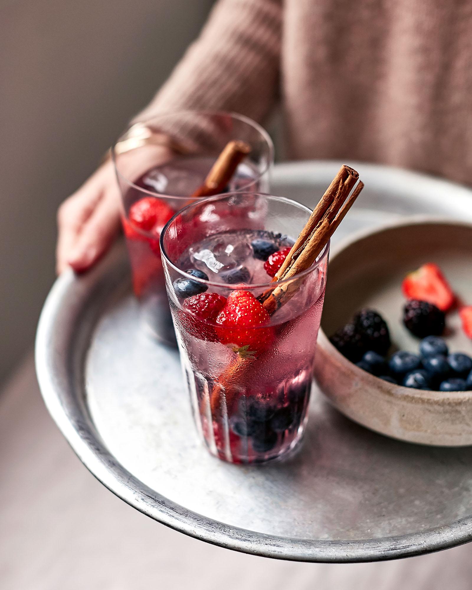 A person holding a tray with two glasses of a pink, fruity drink, garnished with a cinnamon stick and a few berries, on a table with a fruit bowl in the background.