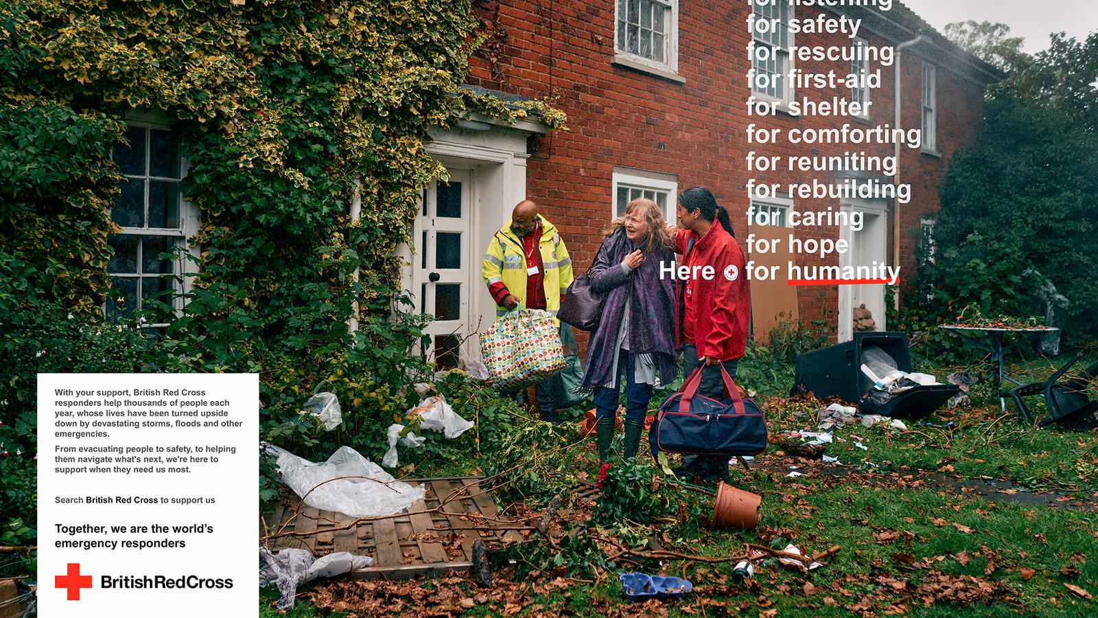 A man in a yellow and red high-visibility jacket holds a large gift-wrapped package in front of a red brick house surrounded by debris.