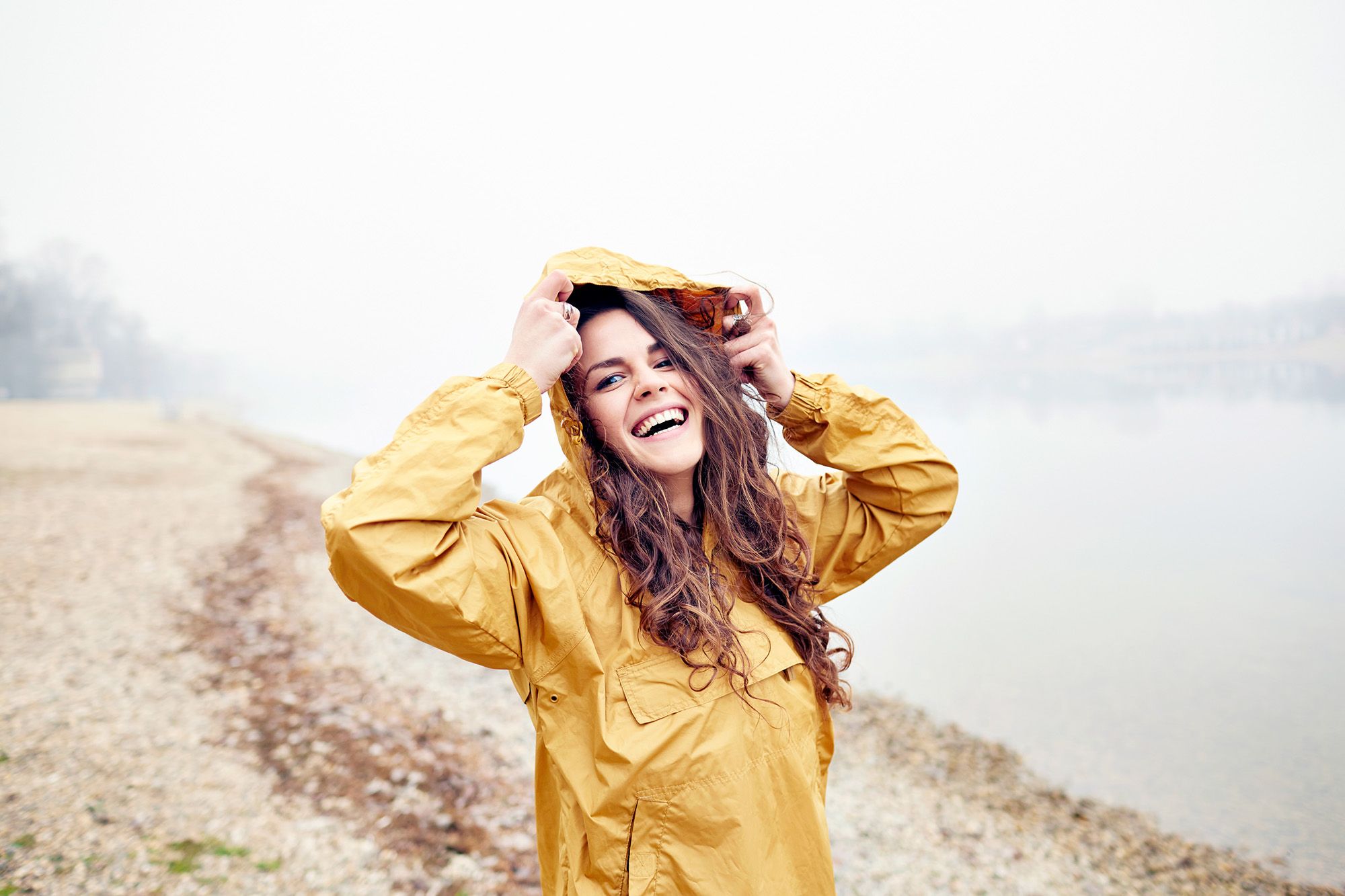 A woman with long, curly brown hair and a bright yellow raincoat, standing in a rainy landscape with a blurred background.