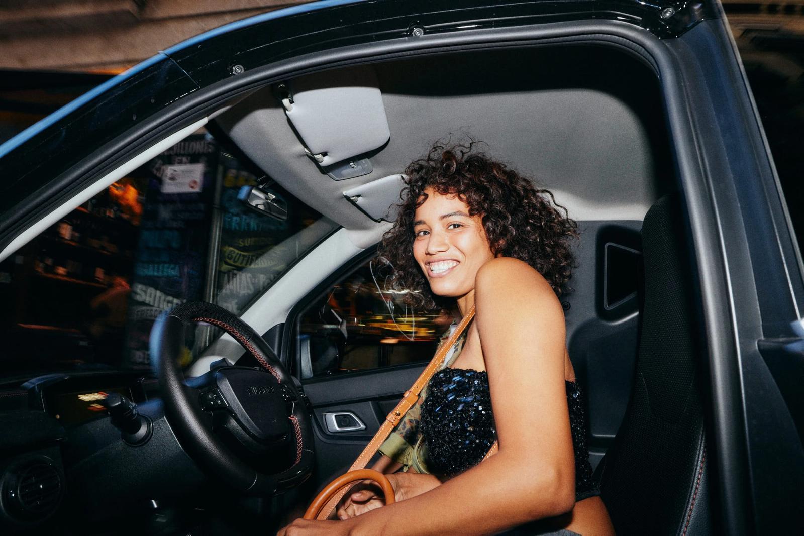 A woman with curly hair and a black sequin top sits in the driver's seat of a car, smiling at the camera with a relaxed, casual expression.