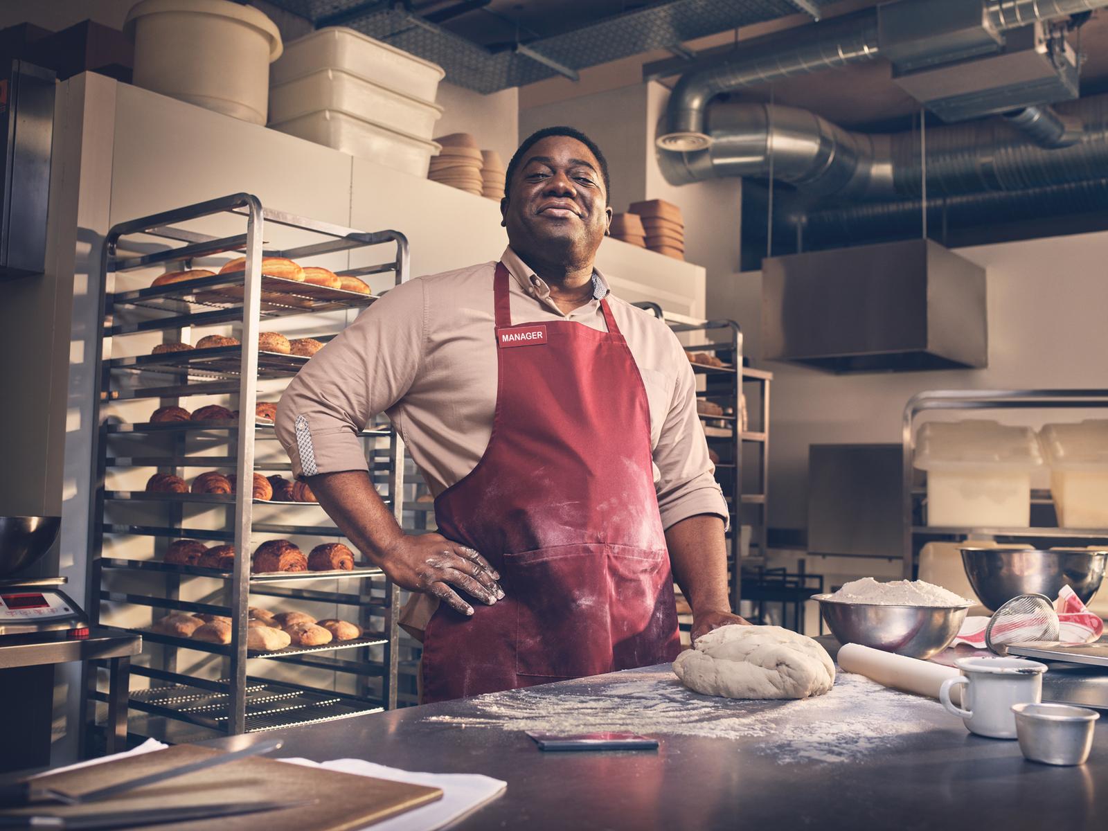 A man in a chef's whites and a red apron stands in a commercial kitchen, surrounded by baking equipment and rows of baked goods on shelves.