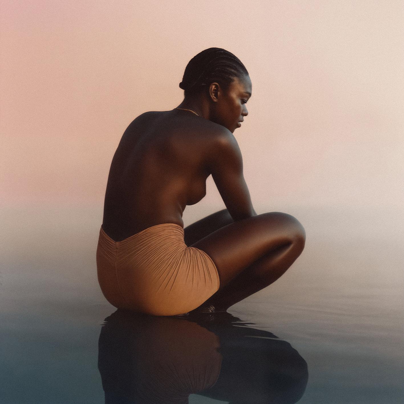 A woman sitting in a lotus position in a body of water, wearing a gold necklace and a pair of tan, high-waisted, pleated shorts.