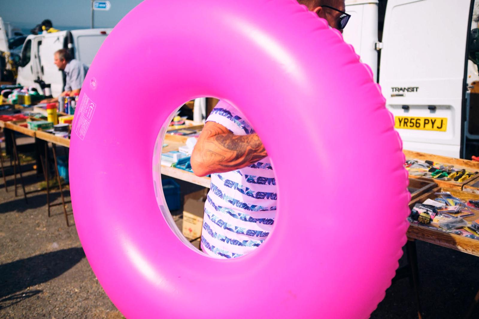 A person with a pink inflatable ring in a busy outdoor market setting. The person is partially obscured by the ring, which is large and bright pink. The person appears to be.