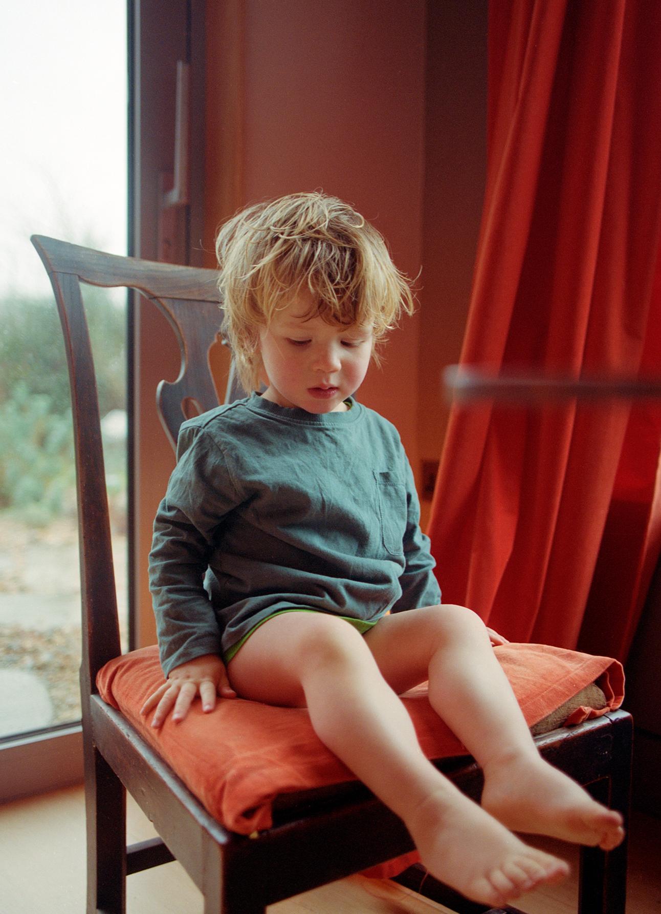 A young child, likely a toddler, is sitting on a chair, wearing a green shirt and no pants.