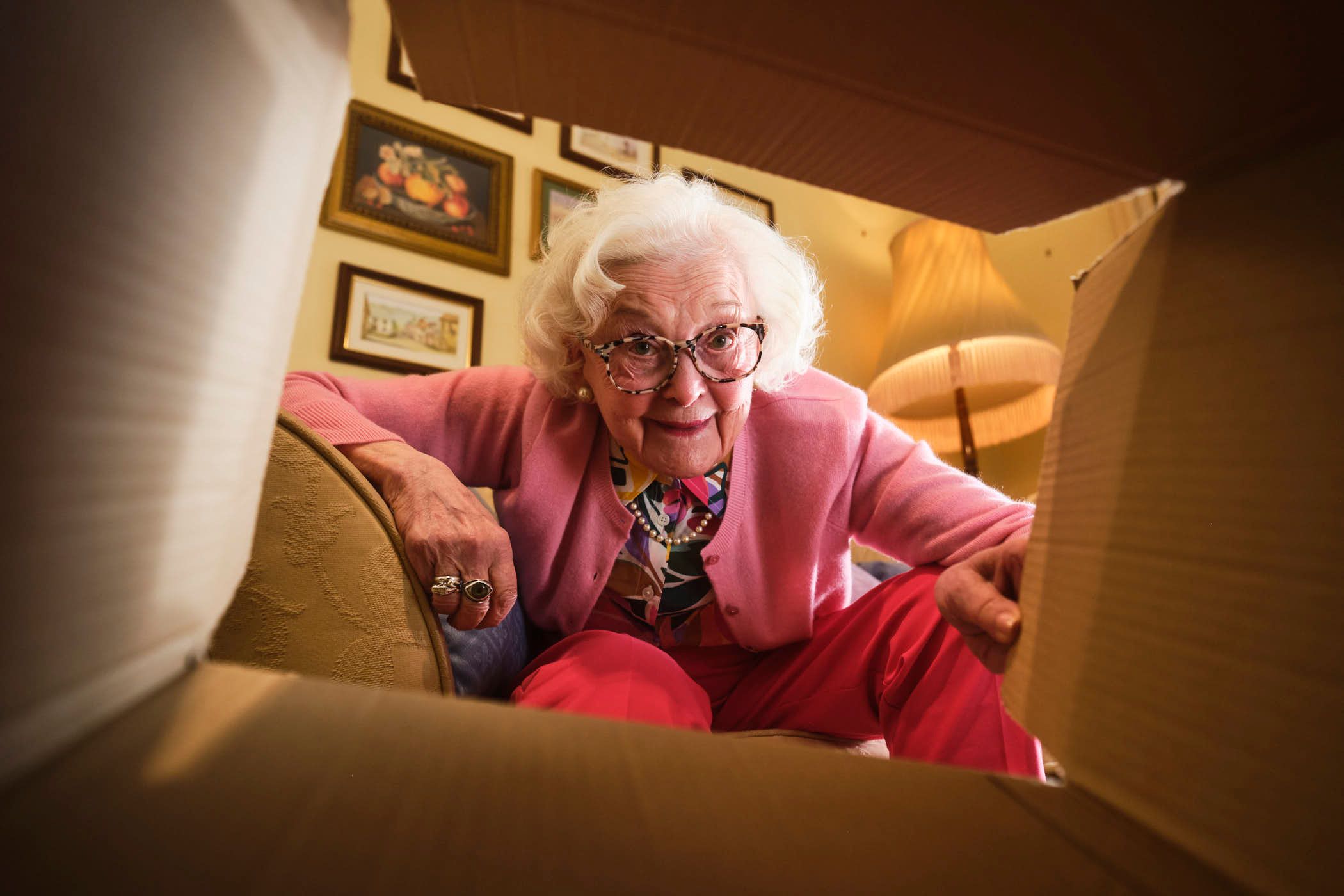 An elderly woman with white hair and glasses is opening a cardboard box, looking directly at the camera with a surprised expression. The woman is dressed in a pink cardigan over.