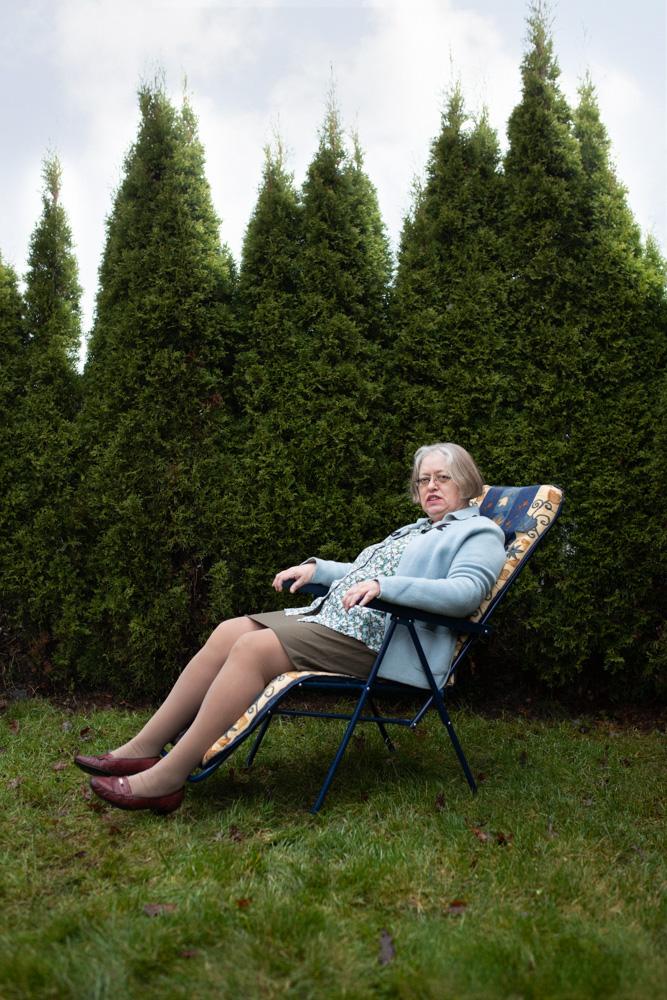 An elderly woman sitting in a chair in a garden, wearing a blue cardigan, a floral blouse, a brown skirt, and brown shoes, with a garden hedge in the background.
