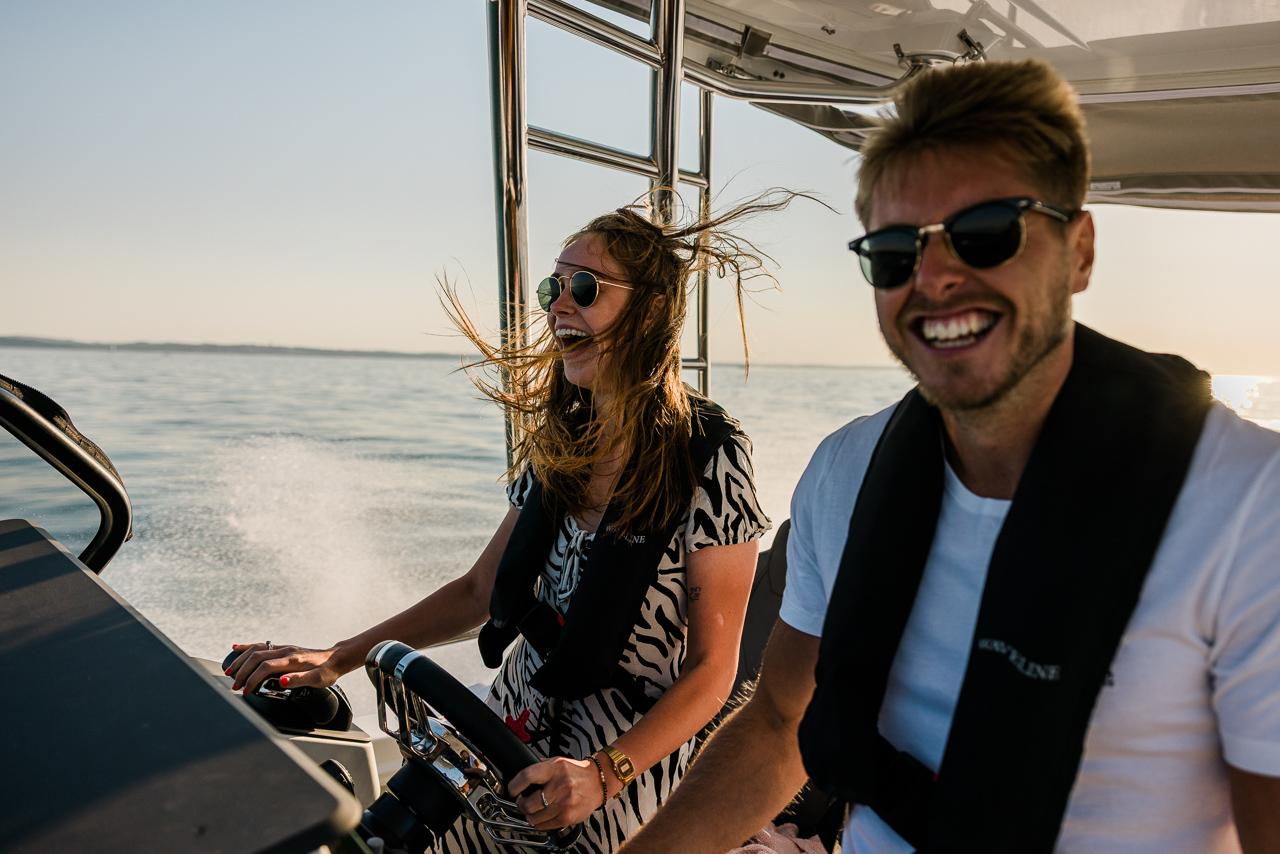 A man and woman are smiling and driving a speedboat on a calm sea.