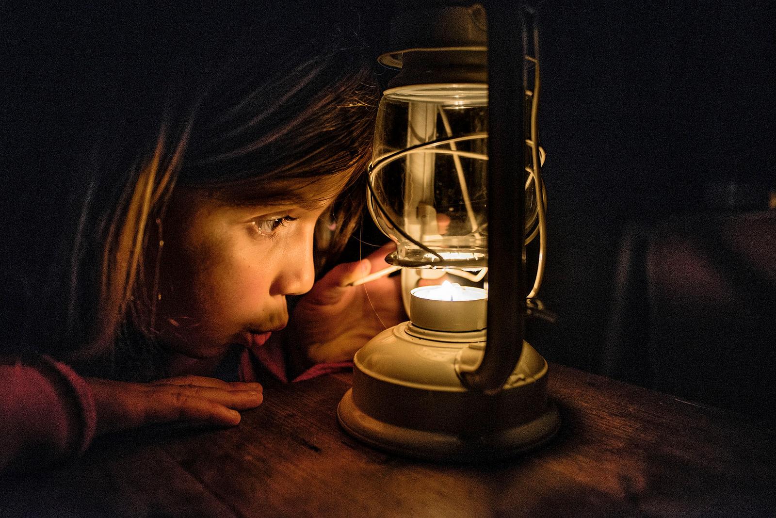 A young girl is blowing out a candle on a lantern, likely in a dark or dimly lit room, with her face illuminated by the warm glow of the flame.