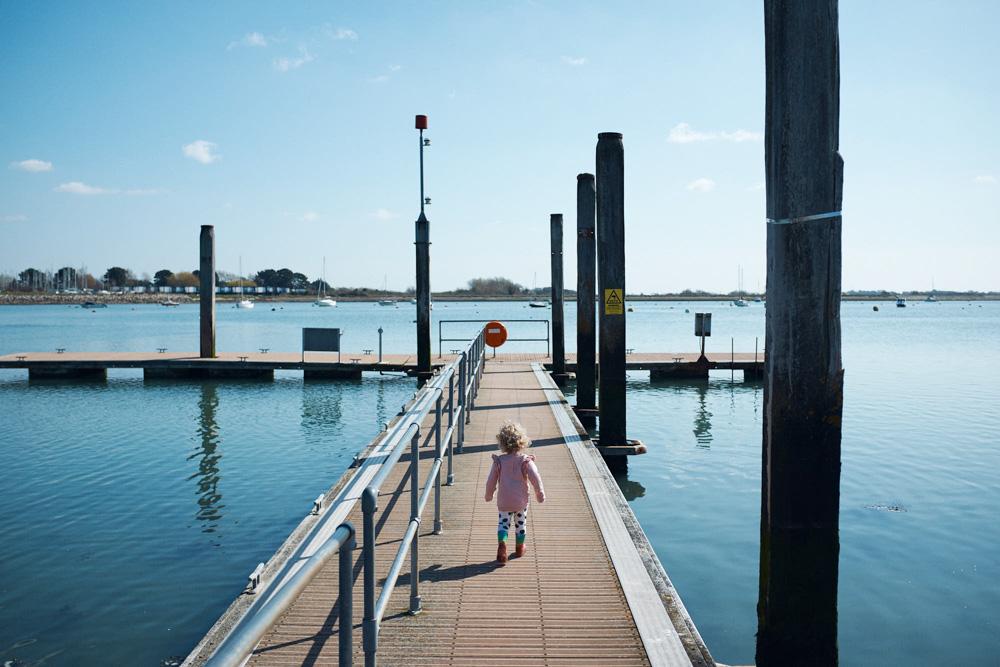 A young girl with curly blonde hair walks alone on a long, narrow dock jutting out into a calm, blue lake. The girl's pink jacket and leggings contrast with the calm, blue water.