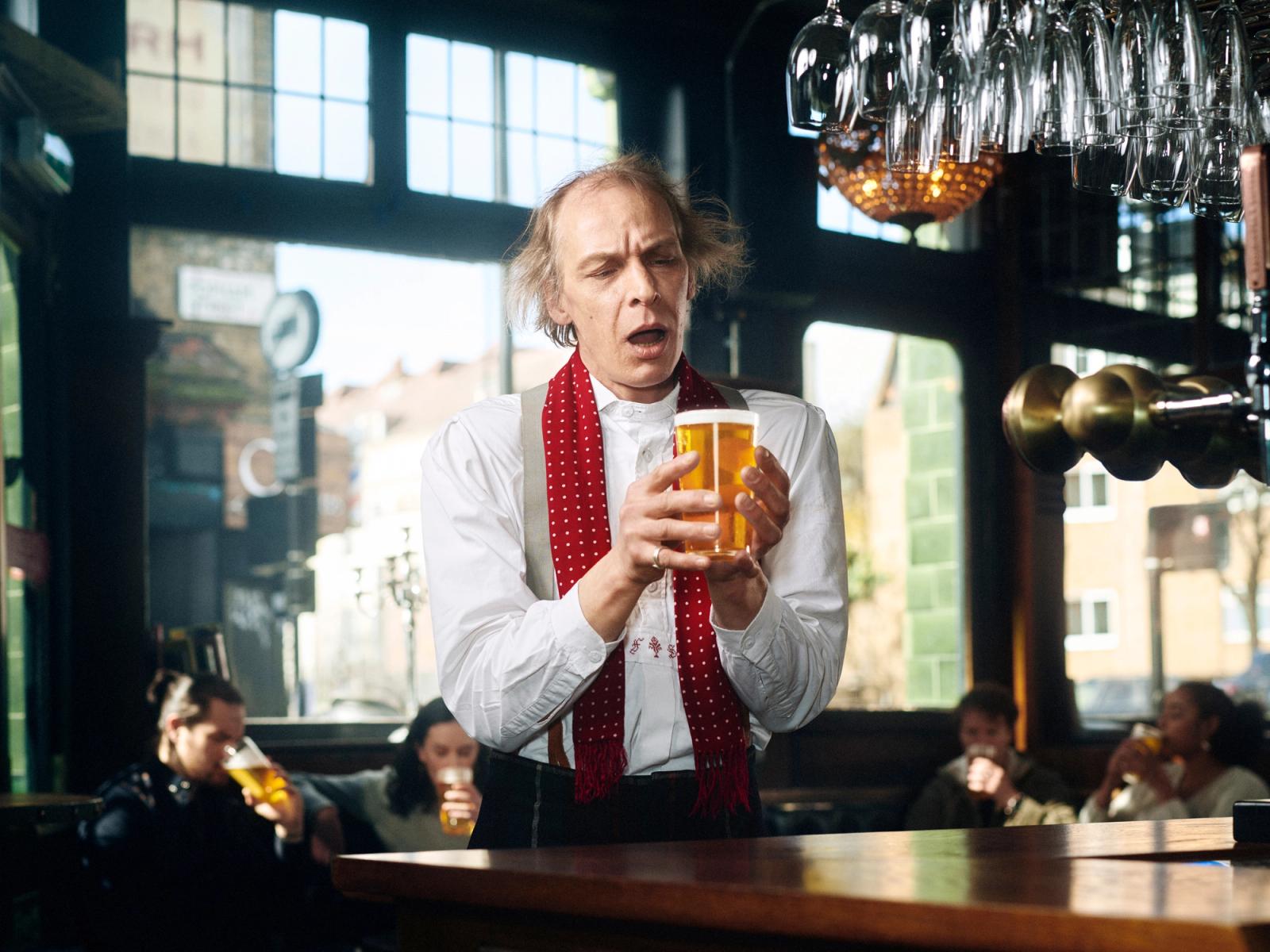 A man in a white shirt, red scarf, and black trousers stands behind a bar, holding a pint of beer.